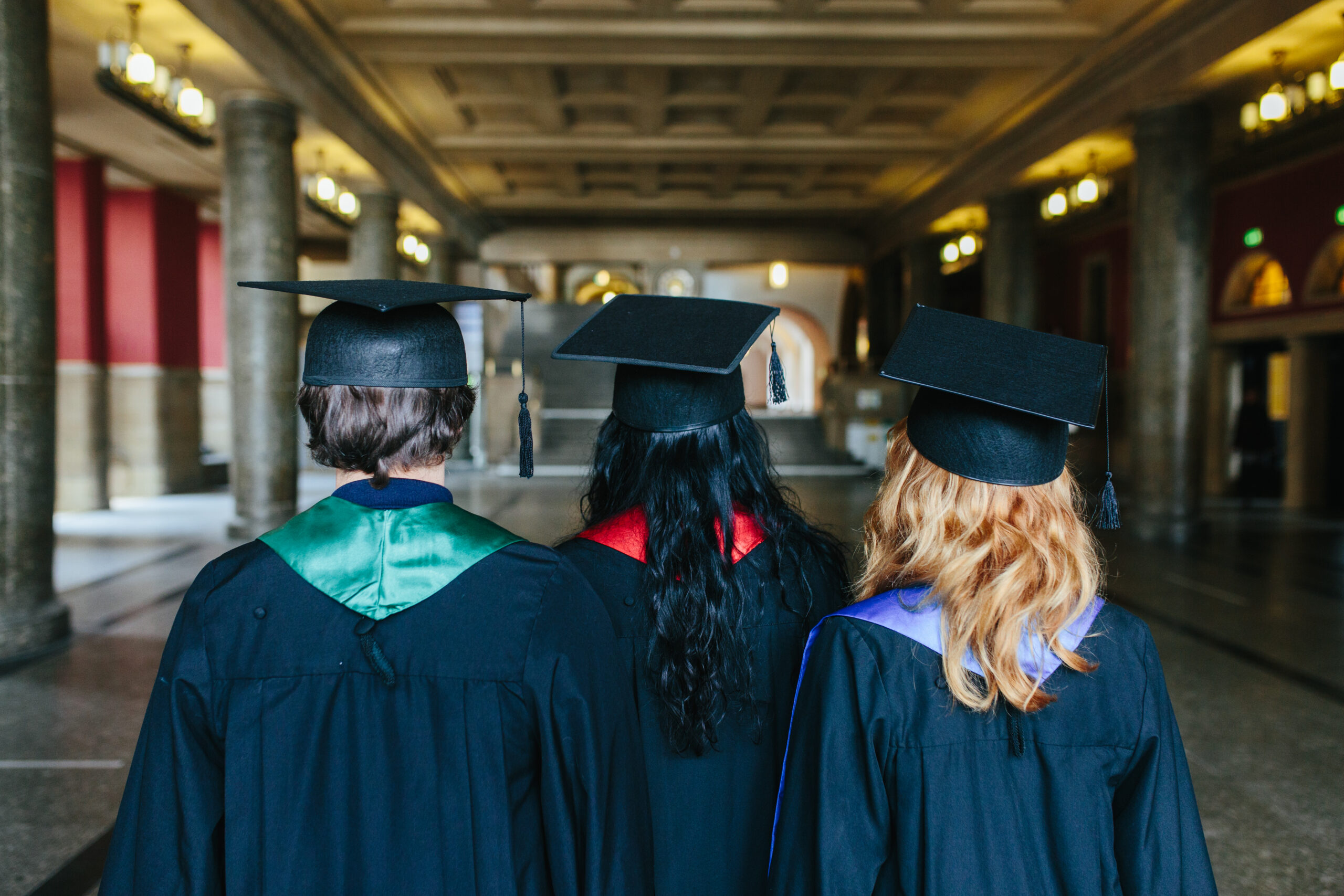 Drei Menschen stehen mit dem Ruecken zur fotografierenden Person und blicken in die Eingangshalle des KG1 der Uni Freiburg. Sie tragen schwarze Umhaenge, sogenannte Talare und schwarze viereckige Huete mit einer Quaste, die sie als Alumni kenntnlich machen