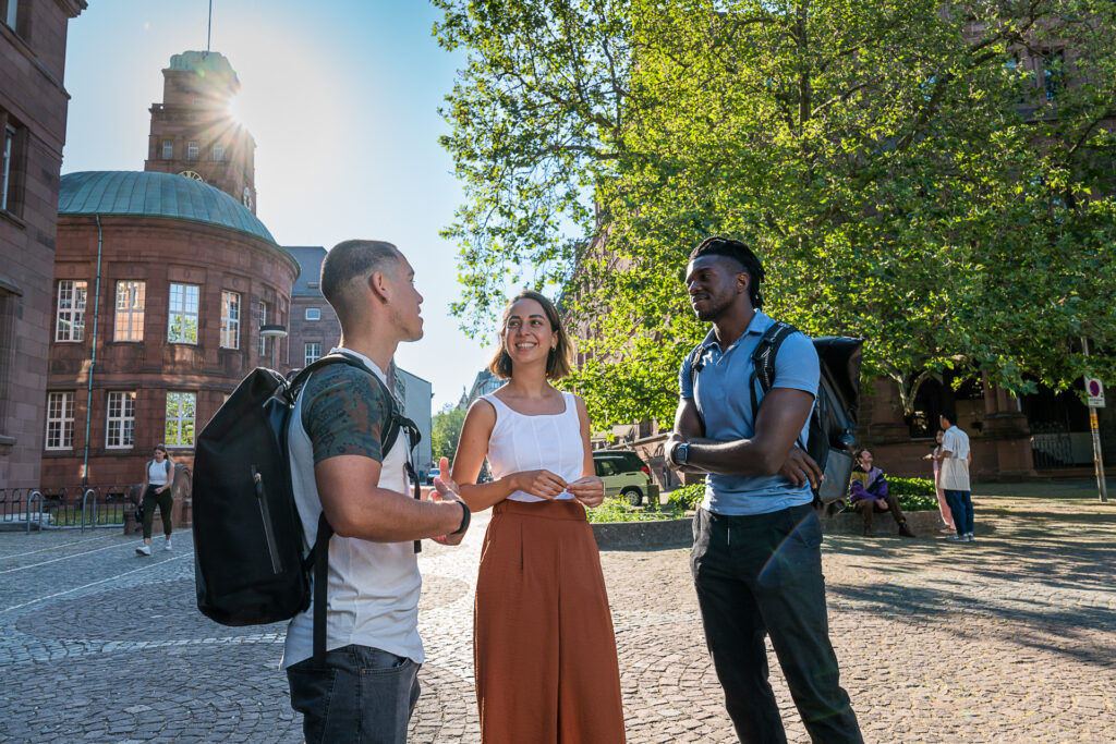 Drei junge Menschen unterhalten sich lebhaft vor einem Universitaetsgebaude der Uni Freiburg