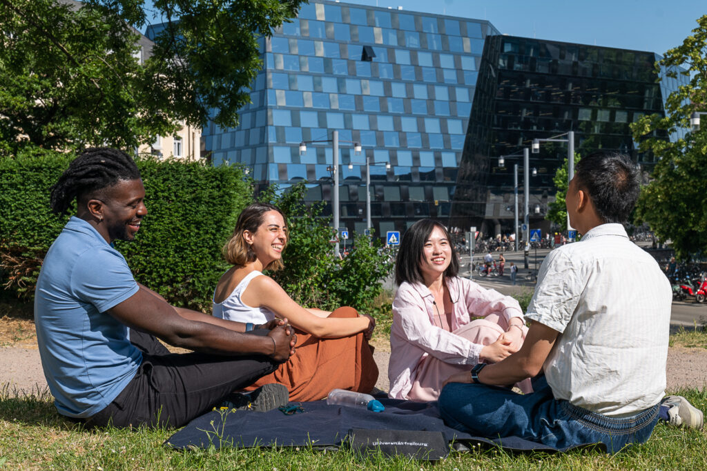 Vier junge Menschen sitzen auf einer Wiese neben der Mensa und lachen und unterhalten sich. Im Hintergrund ist die Universitaetsbiblitohek der Uni Freiburg zu sehen