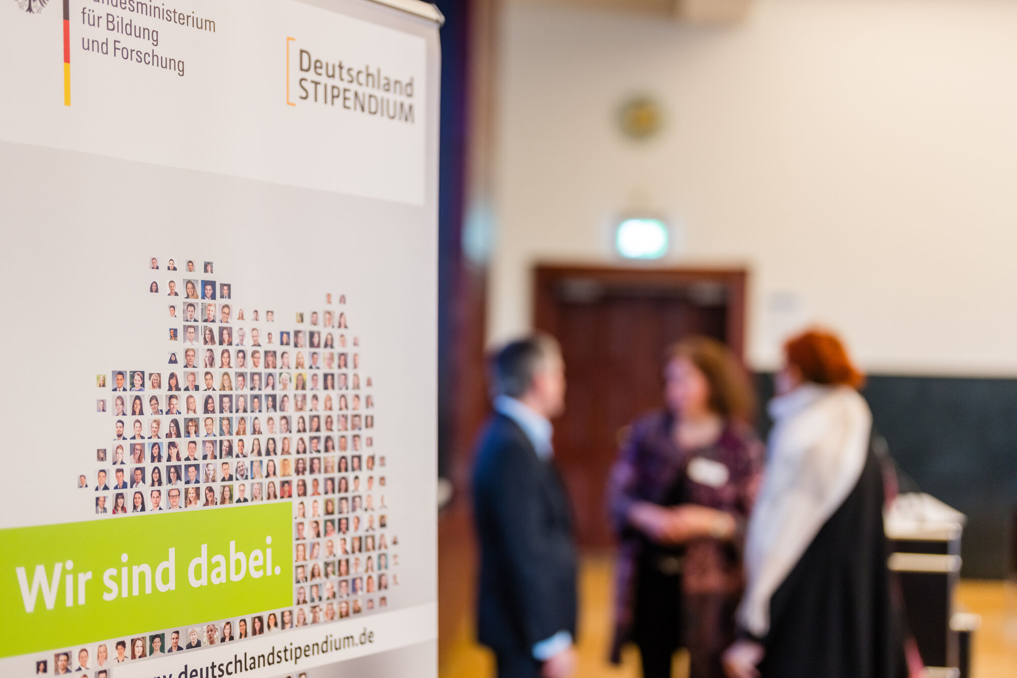 In the foreground is a poster about the Germany Scholarship with the slogan ‘We're in’. A group of people talking to each other can be seen in the background.