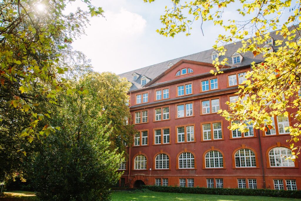 Ein vierstöckiges rotes Gebäude bei Sonnenschein, eingerahmt von grünen Bäumen und Büschen.