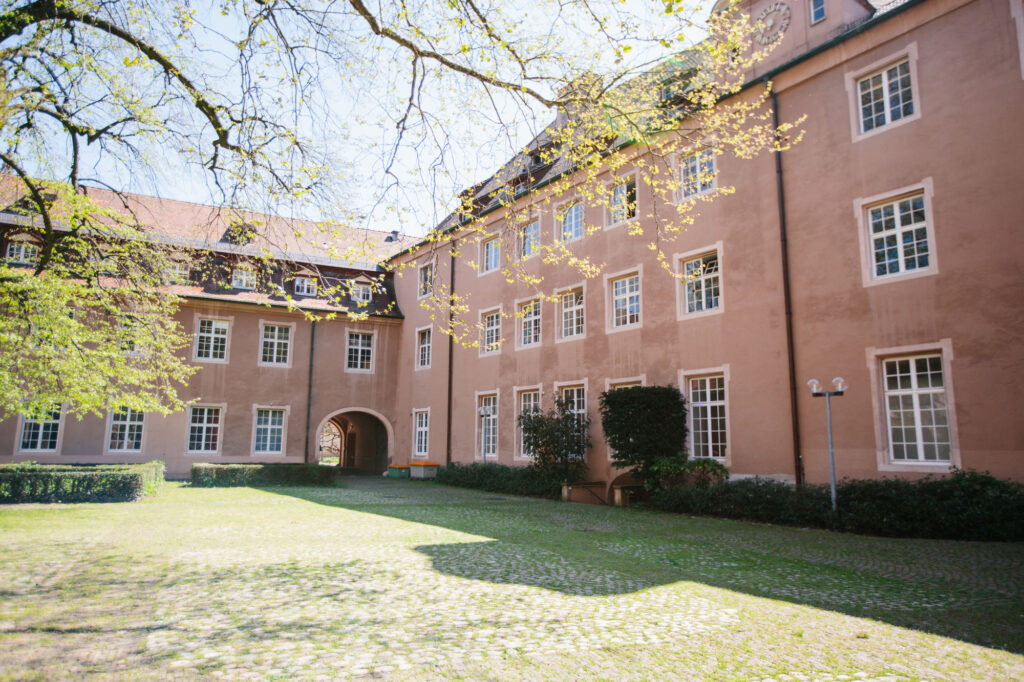 An inner courtyard in spring, surrounded by a red three-storey house. It is the inner courtyard of the so-called Old University of Fribourg, where the UCF University College Fribourg is located.