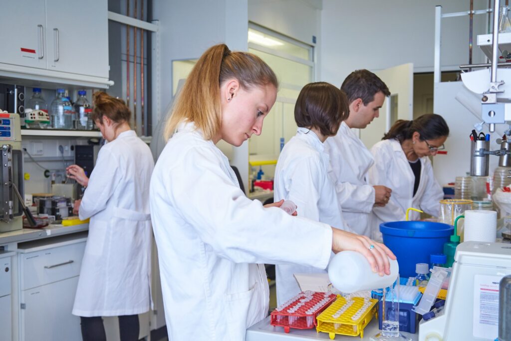 Five young researchers are working in a laboratory. They are wearing white coats. The researcher at the front of the picture is filling a liquid into a cylinder.