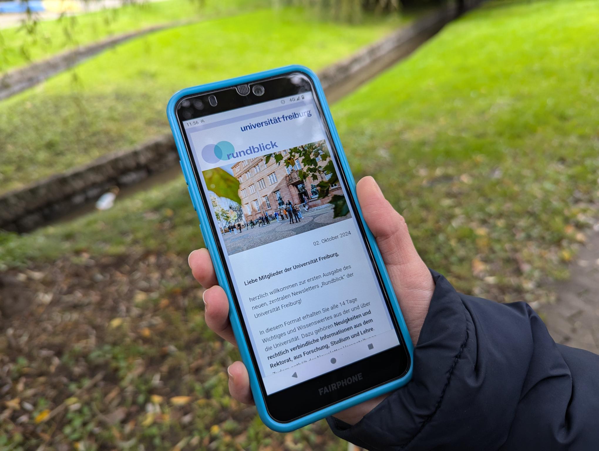 A hand holds a smartphone in front of a green background. The smartphone shows the editorial of the ‘Rundblick’ newsletter.