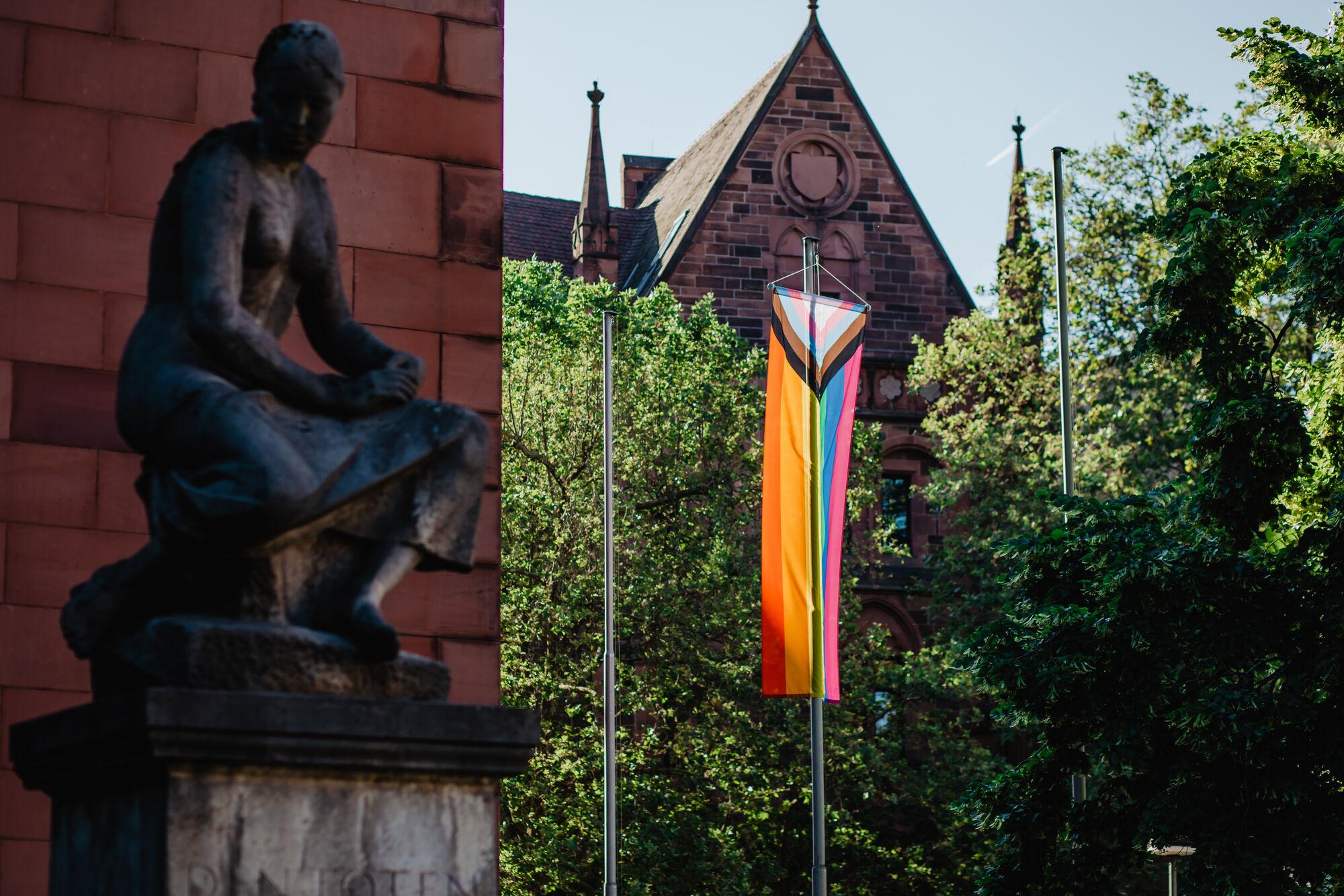 A rainbow flag in front of old buildings at the University of Freiburg. A statue of a woman can be seen in the foreground.