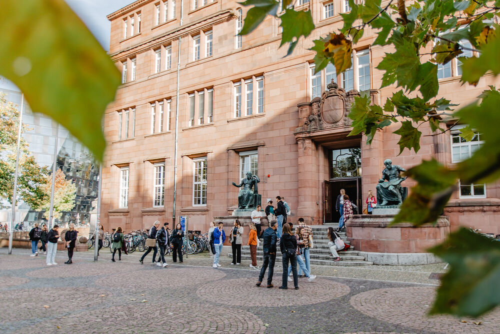 The Kollegiengebäude I of the University of Freiburg with people in the front. Also, autumn leaves are in the forefront of the picture.