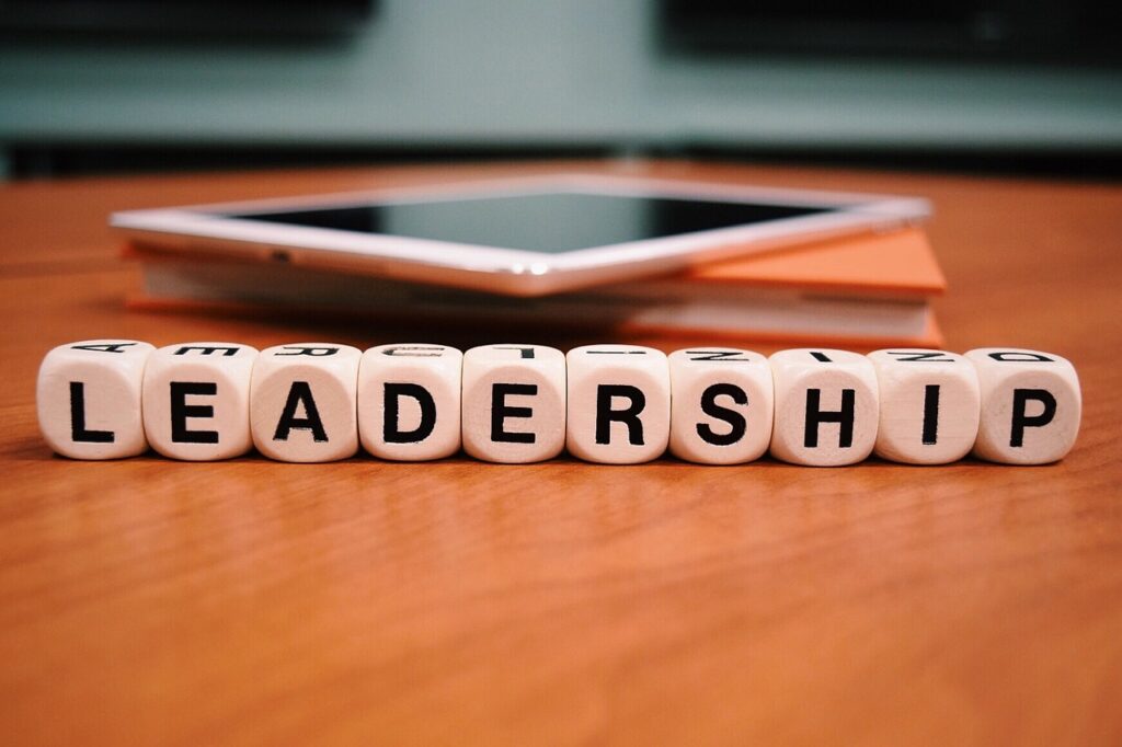 Dice forming the word "Leadership". The dice are on a wooden table, in the background one sees a tablet and a notebook