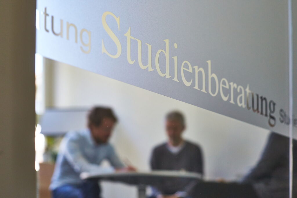 Zwei Personen im Beratungsgespräch hinter einer Glaswand mit der Aufschrift Studienberatung