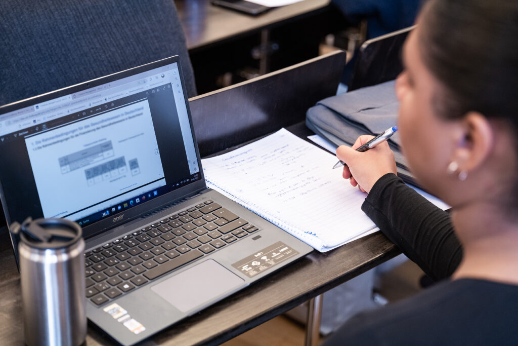 Blick im Hörsaal auf den Laptop und die Notizen einer Studentin