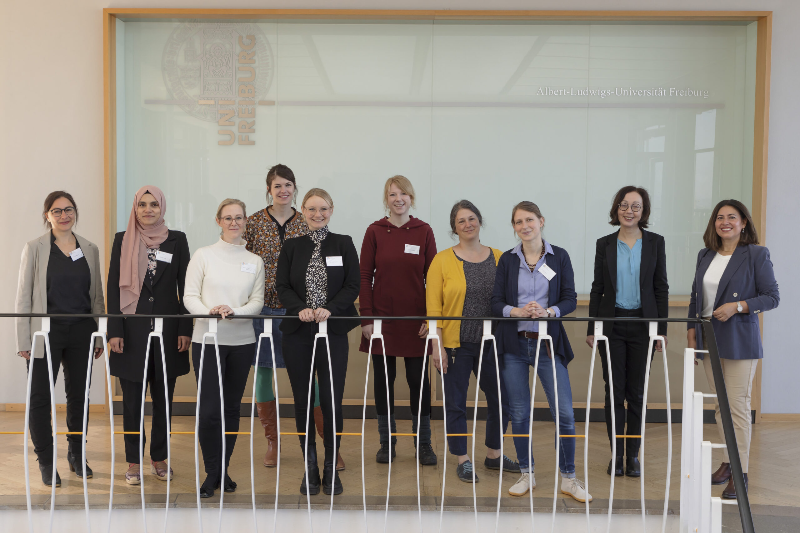 Eight program participants, the Vice-Rector, and the program director are standing smiling in front of a glass wall displaying the university logo.