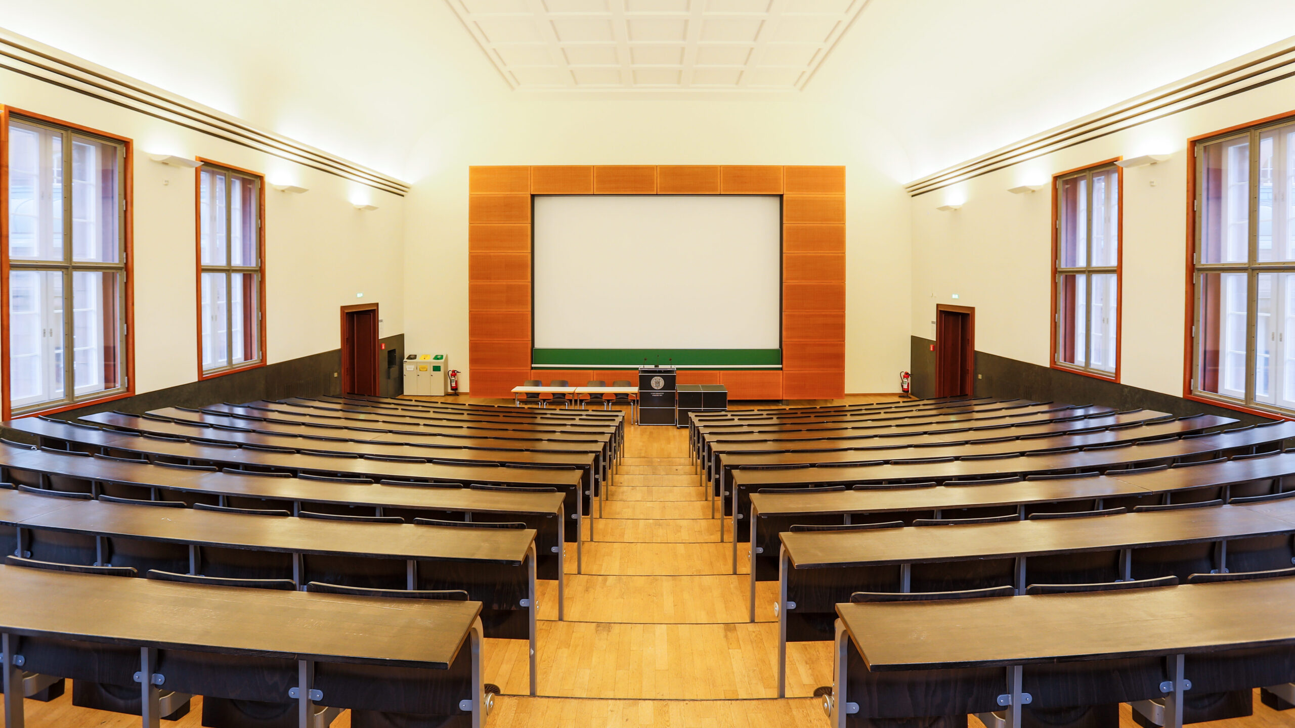 Ein leerer Hörsaal, der Universität Freiburg. Es laufen zwei Sitzreihen auf das Sprechpult zu. Zwischen den Sitzreihen befindet sich ein Mittelgang. Hinter dem Sprechpult ist eine Leindwand angebracht.