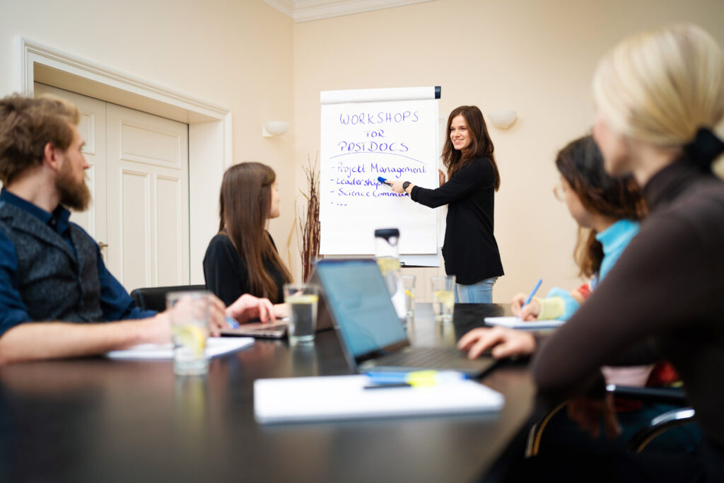 A woman stands at a flipchart and explains a topic to a group of listeners. The flipchart sheet reads Workshops for Postdocs.