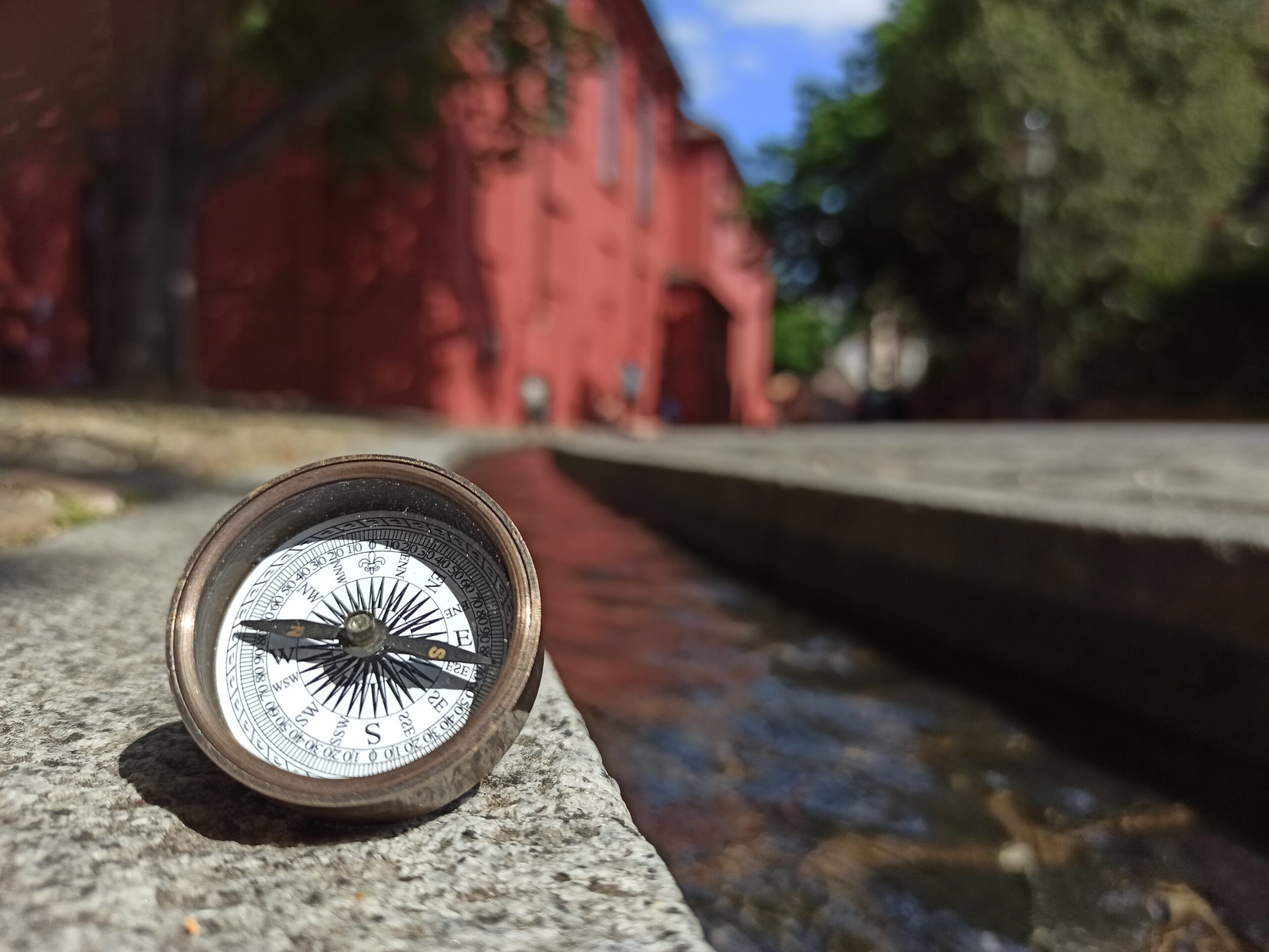 An old compass can be seen in the foreground on the left. A stream flows past it. A red building and trees can be seen in the background.