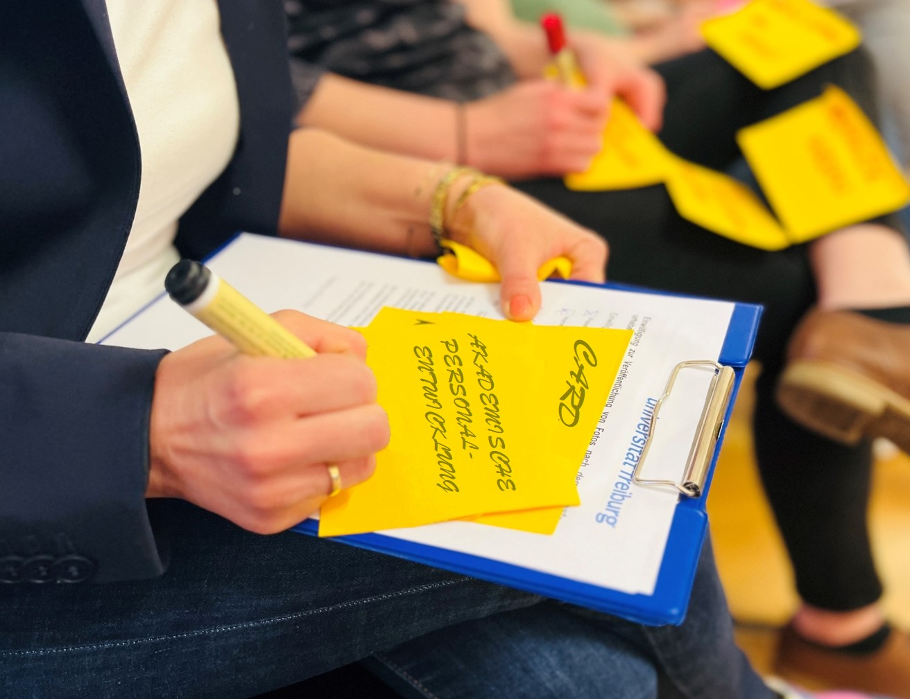 Eine Frau hält ein Klemmbrett in der Hand und schreibt mit einem schwarzen Stift auf einen gelben Zettel. Auf dem Zettel ist zu lesen "CARD" und "Akademische Personalentwicklung".