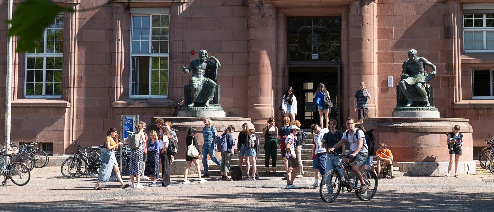 Eine größere Grupe von Studierenden der Universität Freiburg vor dem Eingang des KG 1, die das Gebäude betreten oder verlassen. Links und rechts vom Eingang sind Bronzefiguren der Philosophen Homer und Aristoteles.