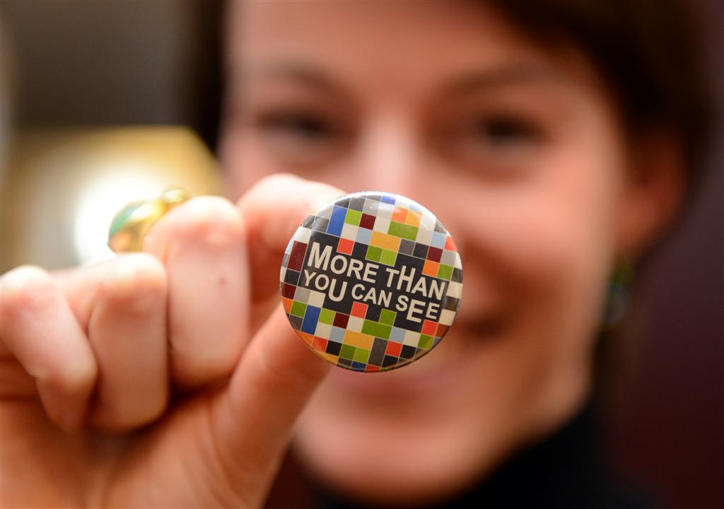 Eine lächelnde Frau hält einen Button zum Anstecken in die Kamera. Auf dem Button steht: More than you can see. Die Frau trägt einen goldenen Ring mit einem türkisfarbenen Stein an ihrem linken Mittelfinger.