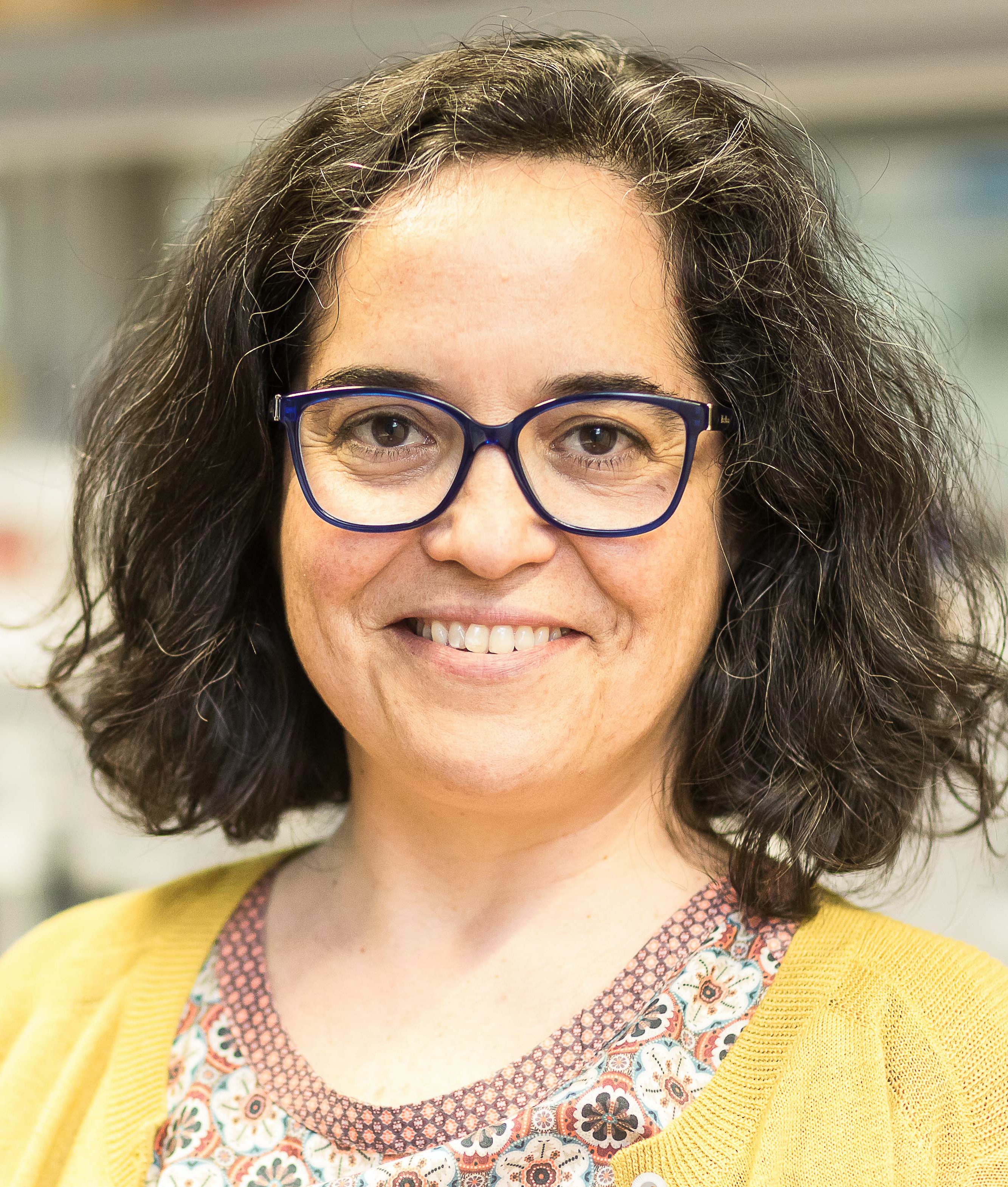 a portrait of Ana Pombo. She is a friendly looking white colored woman with dark blues glasses and curly brown hair to her shoulders.