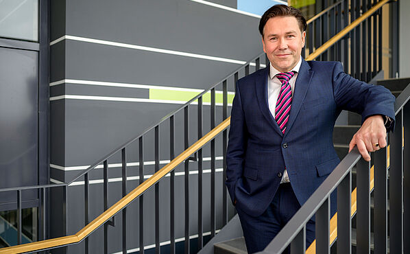 The picture shows Prof. André Fischer. He stands on stairs inside a modern looking building. He wears a dark suit and a red-blue striped tie. He smiles friendly.