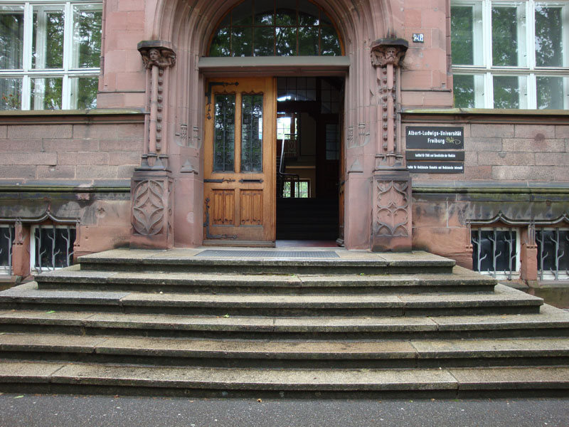 Sechs Treppenstufen empor befindet sich eine alte, große Holztür, neben der ein Schriftzug die Universität ankündigt.