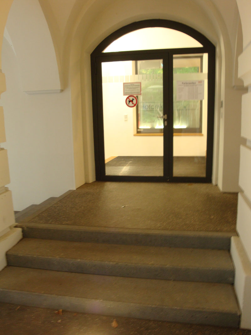 Einige Treppenstufen empor befindet sich eine große Glastür in einem gewölbten Gebäude