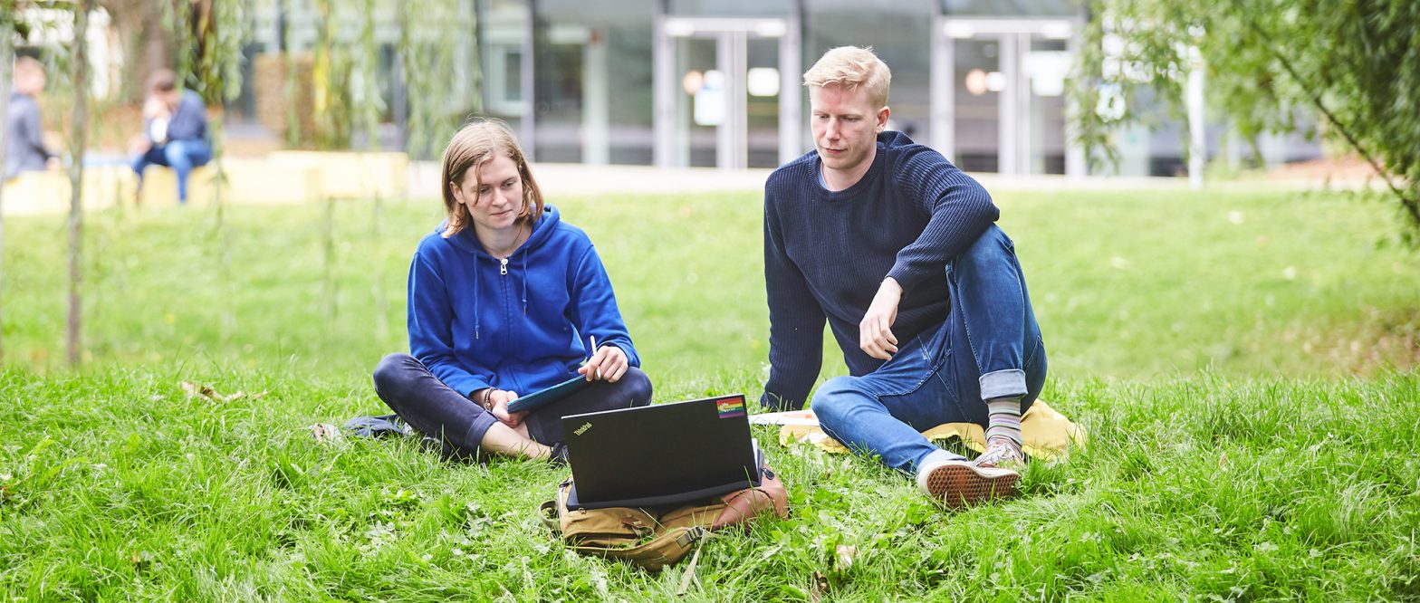 Zwei Studierende sitzen im Grünen mit einem Laptop