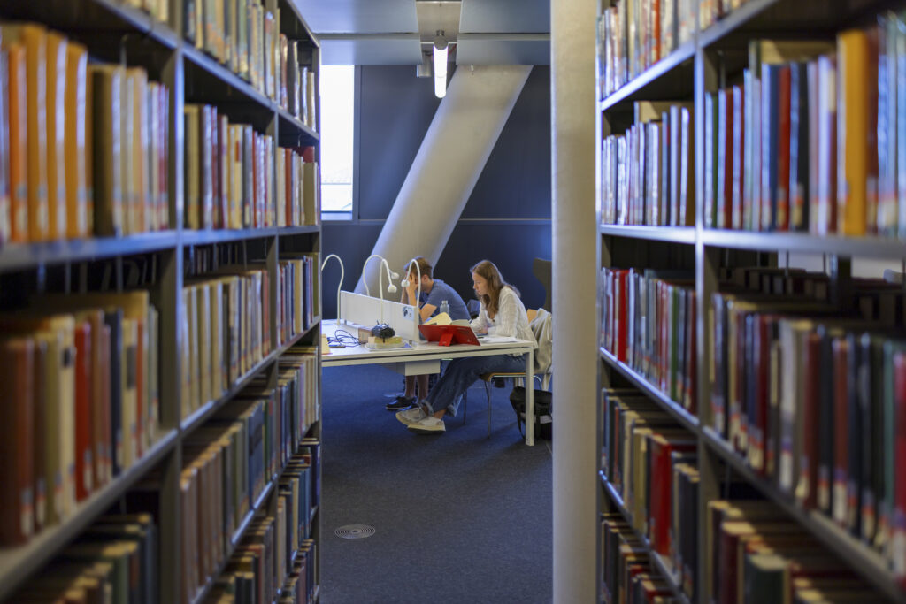 Studierende sitzen in der Bibliothek. Bücher im Vordergrund.