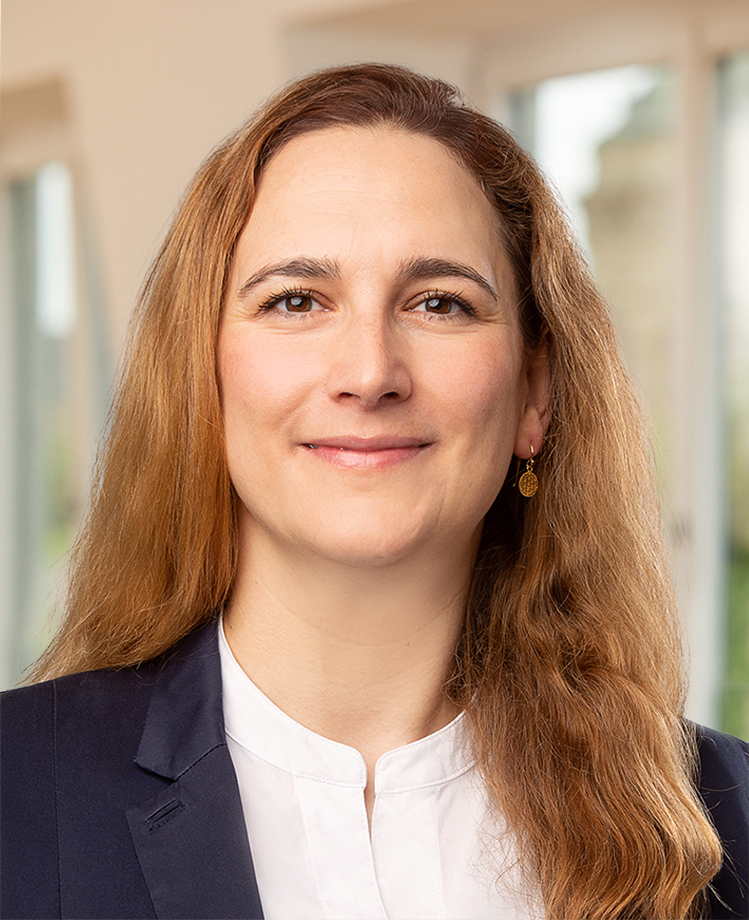 Man sieht das Portraitbild einer Frau mit gewellten, dunkelblonden Haaren und braunen Augen. Sie trägt eine weiße Bluse und einen blauen Blazer. Sie lächelt freundlich in die Kamera.