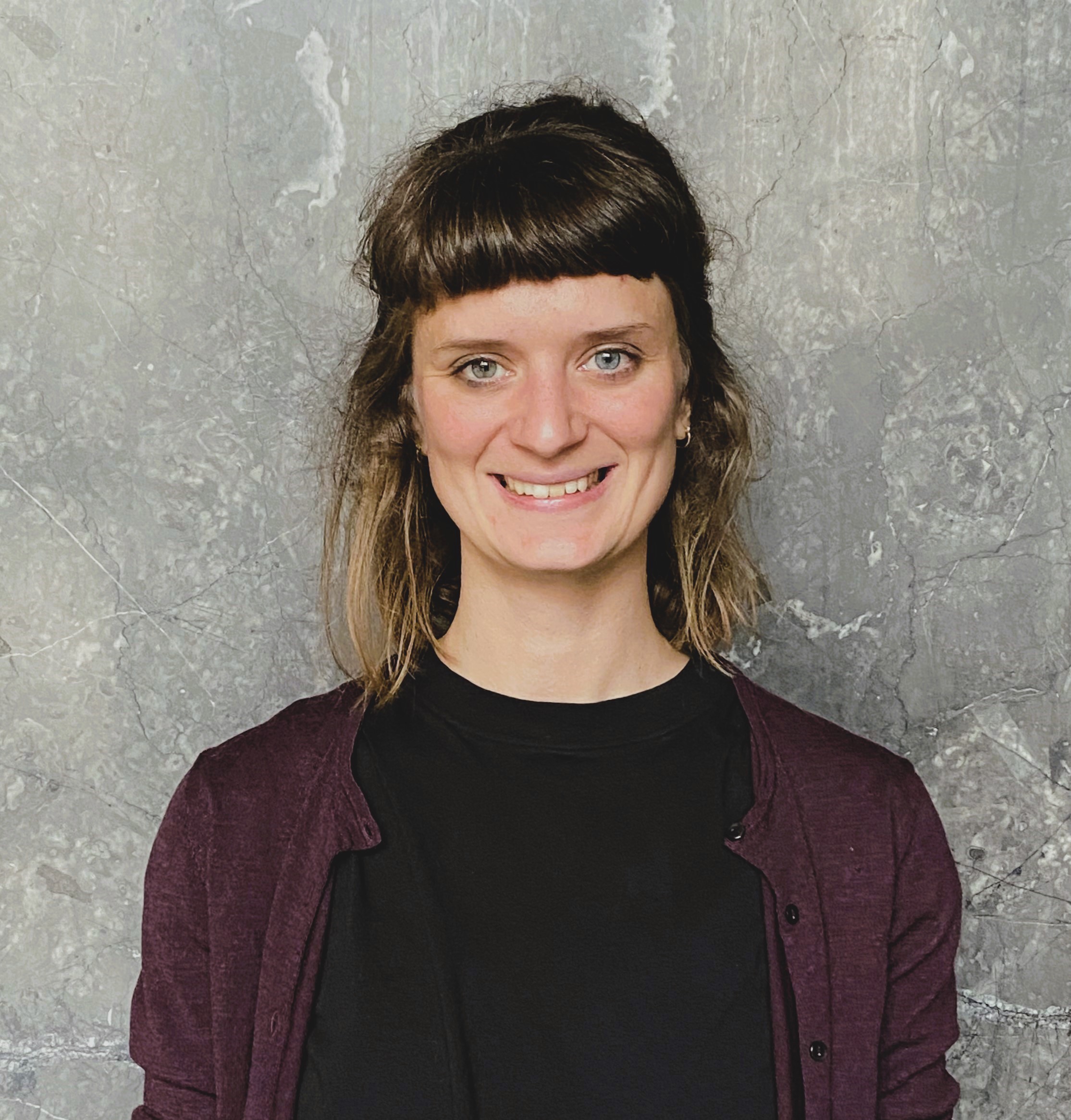 Man sieht das Portraitbild einer Frau mit blauen Augen. Sie hat schulterlanges, leicht gewelltes, braunes Haar mit einem geraden Pony. Sie trägt ein schwarzes T-Shirt und darüber eine lila Strickjacke. Sie lächelt freundlich in die Kamera.