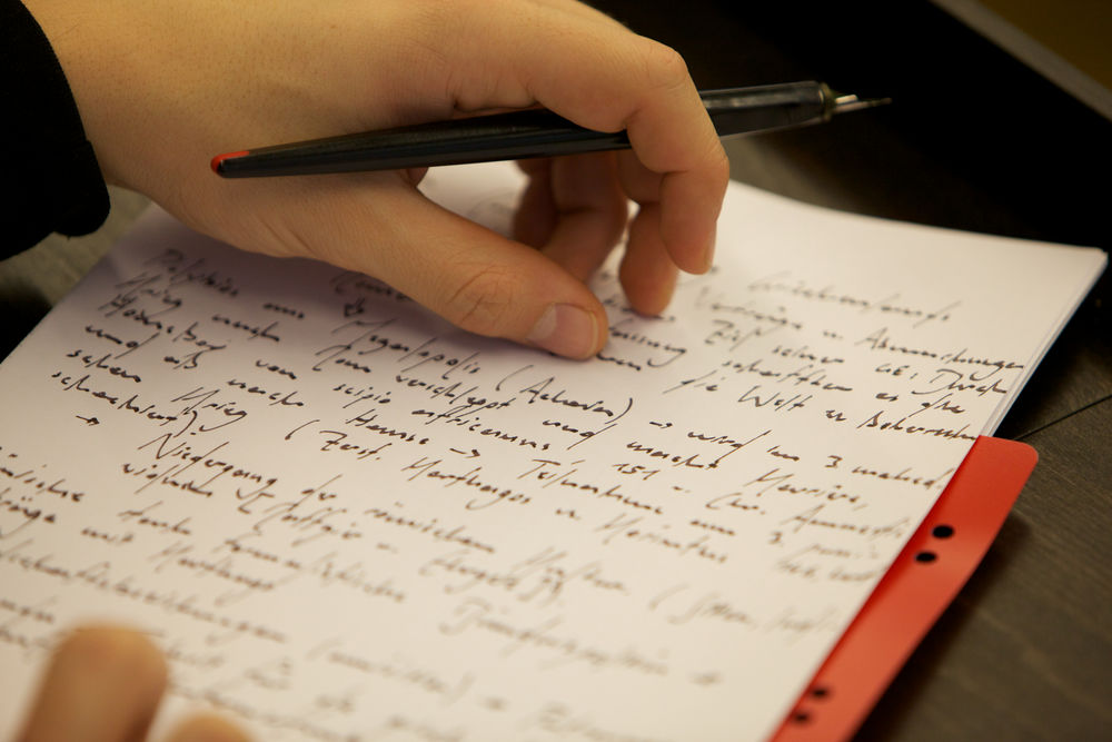 Ein handschriftlich beschriebenes Blatt Papier mit einer Hand, die einen Füllfederhalter hält.