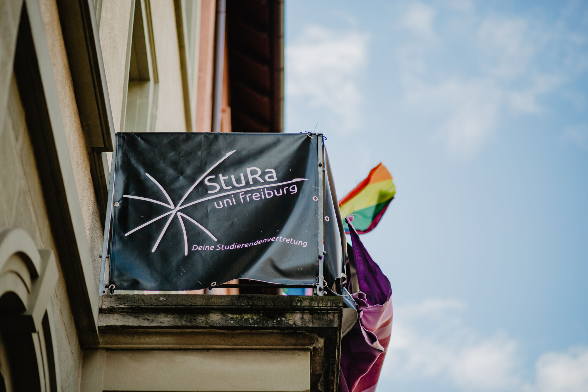 Ein Balkon von unten. Eine Plane mti dem Logo der Verfassten Studierendenschaft (StuRa) der Universität Freiburg sowie eine Regenbogenfahne sind zu erkennen.