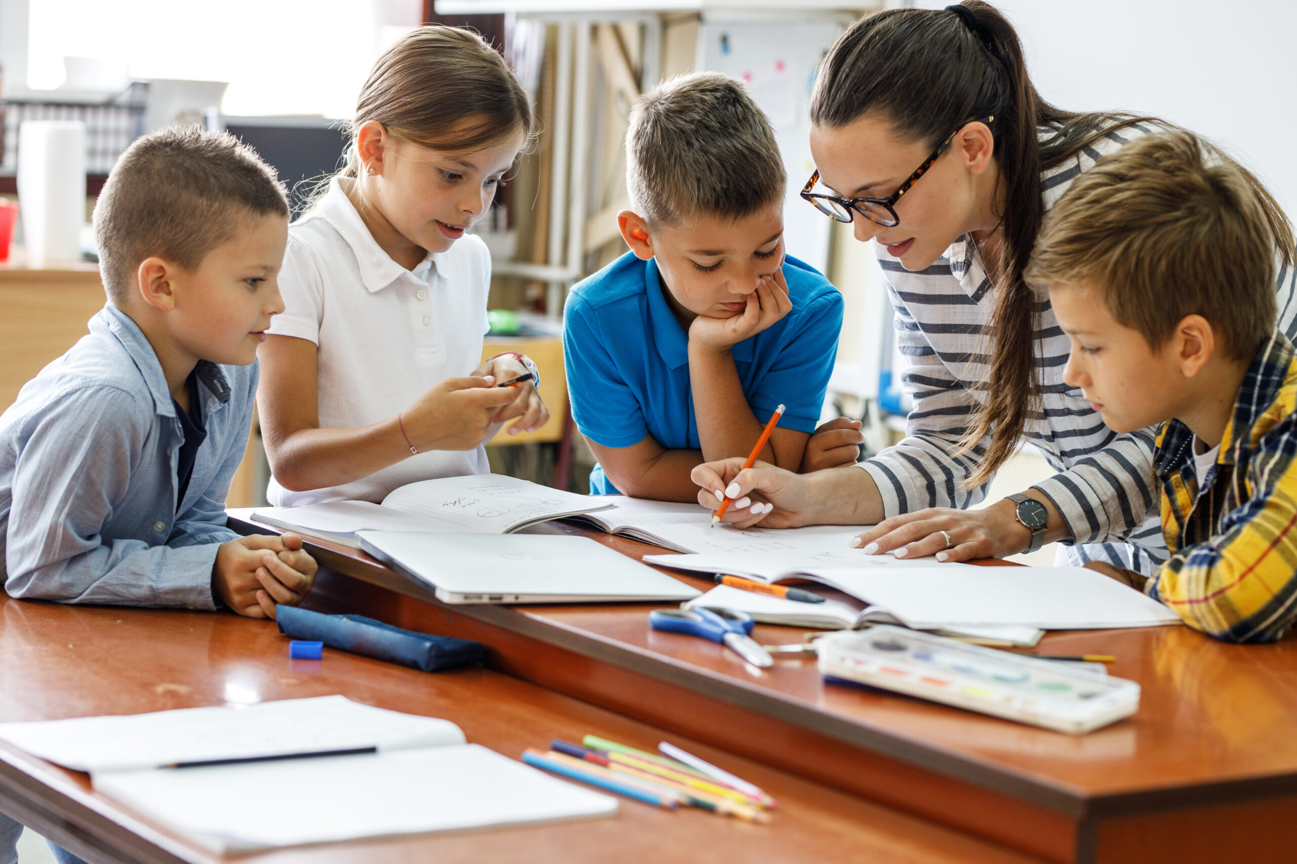 Eine Lehrerin bespricht mit Schulkindern Aufgaben an einem Schreibtisch im Klassenzimmer.