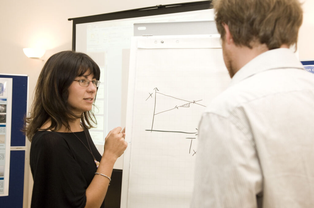 Eine Frau mit Brille und halblangen braunen Haaren steht an einem Flipchart, auf dem ein Schaubild gezeichnet ist, und spricht mit einem Mann, der mit dem Rücken zur Kamera steht.