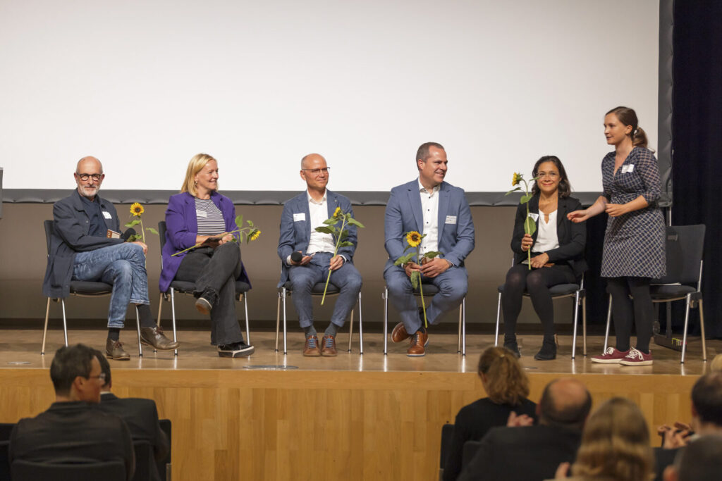 Rednerinnen und Redner sitzen in einer Reihe auf Stühlen auf der Bühne und haben jeweils eine Sonnenblume in der Hand.