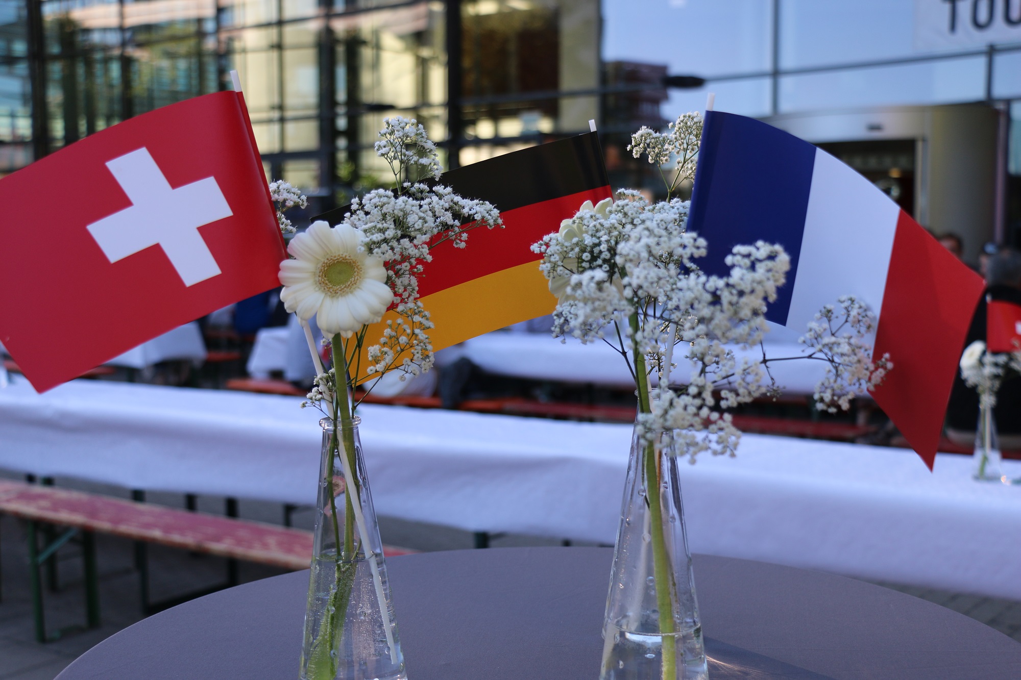 Die Flaggen der Schweiz, Deutschland und Frankreich stehen in zwei Blumenvasen nebeneinander.