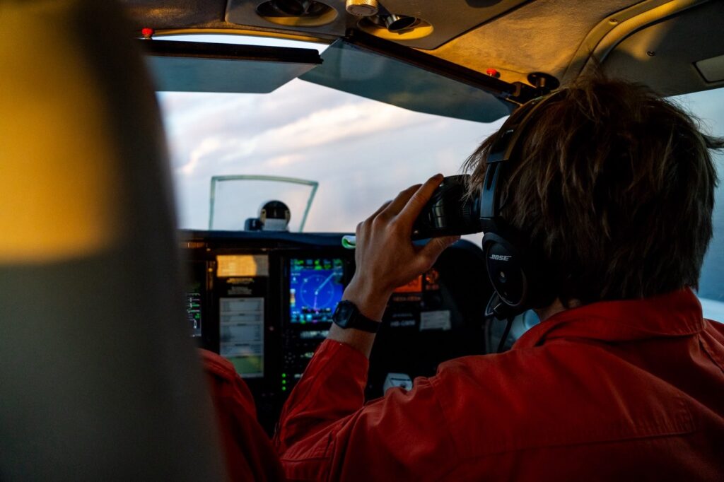 Blick auf ein Flugzeug-Cockpit. Vorne rechts sitzt Joseph Oertel, der durch ein Fernglas schaut.