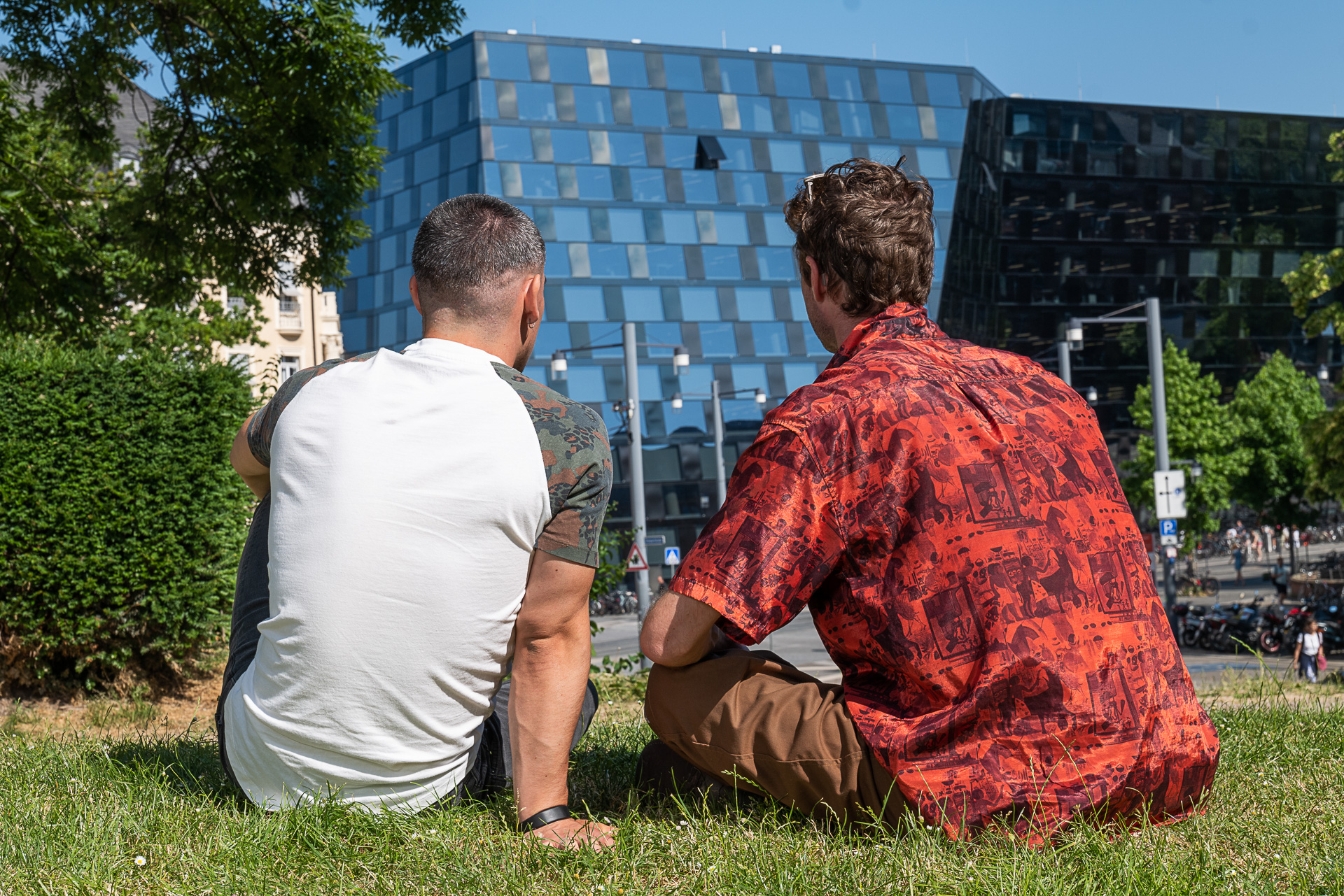 Zwei junge Männer sitzen im Gras, man sieht sie von hinten. Beide blicken auf die Universitätsbibliothek, die man ebenfalls sieht.