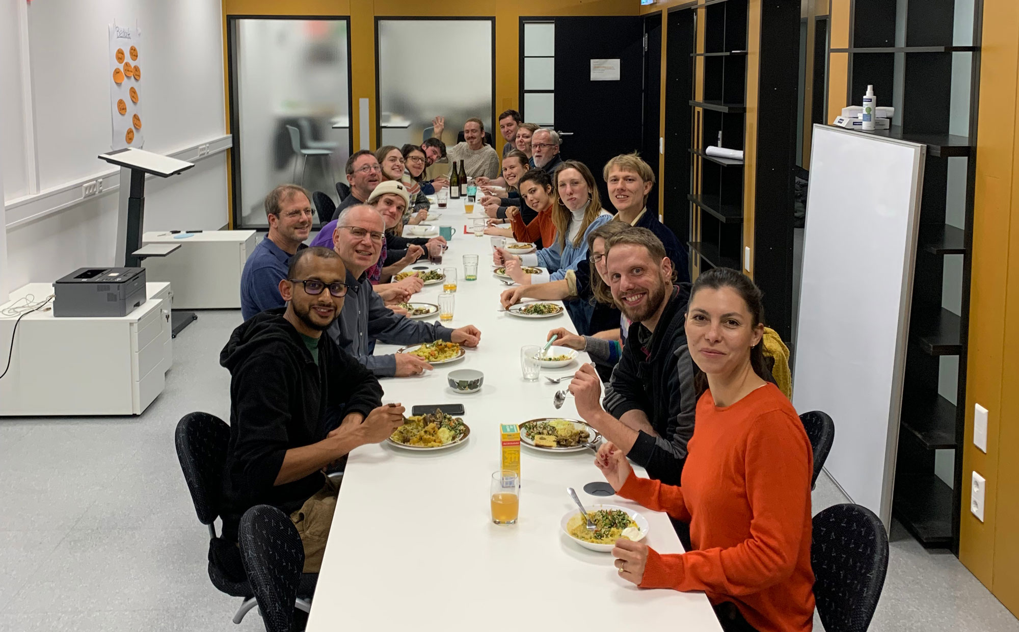 eine Grupe von Menschen an einem langen Tisch mit Essen. Alle schauen in die Kamera.