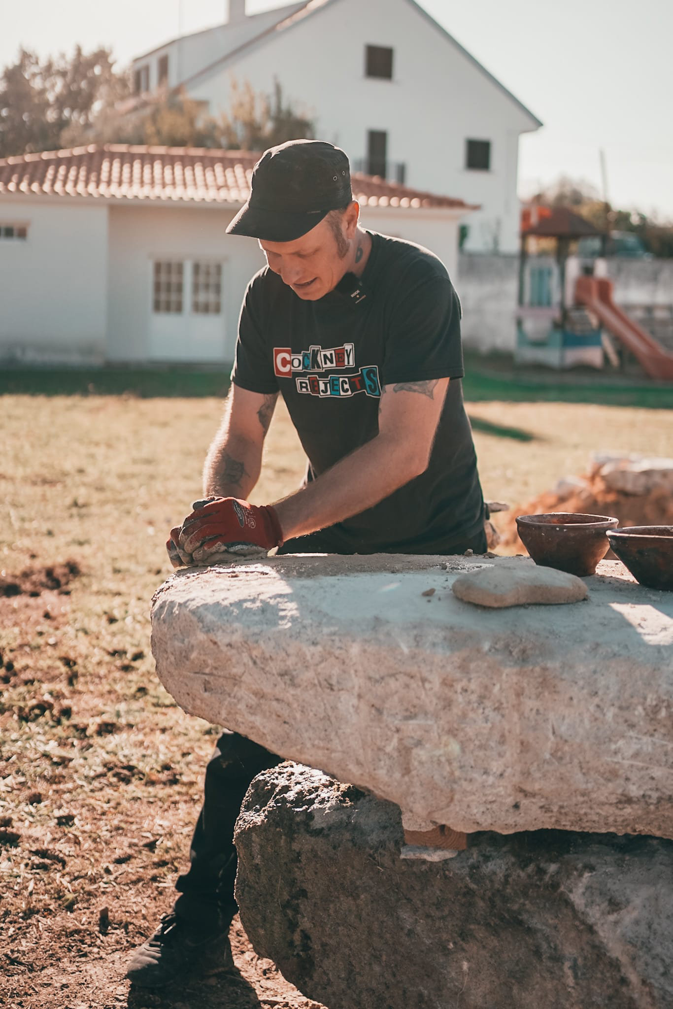 Mann mit Mütze und Handschuhen bearbeitet Stein im Freien, im Hintergrund ein Haus.