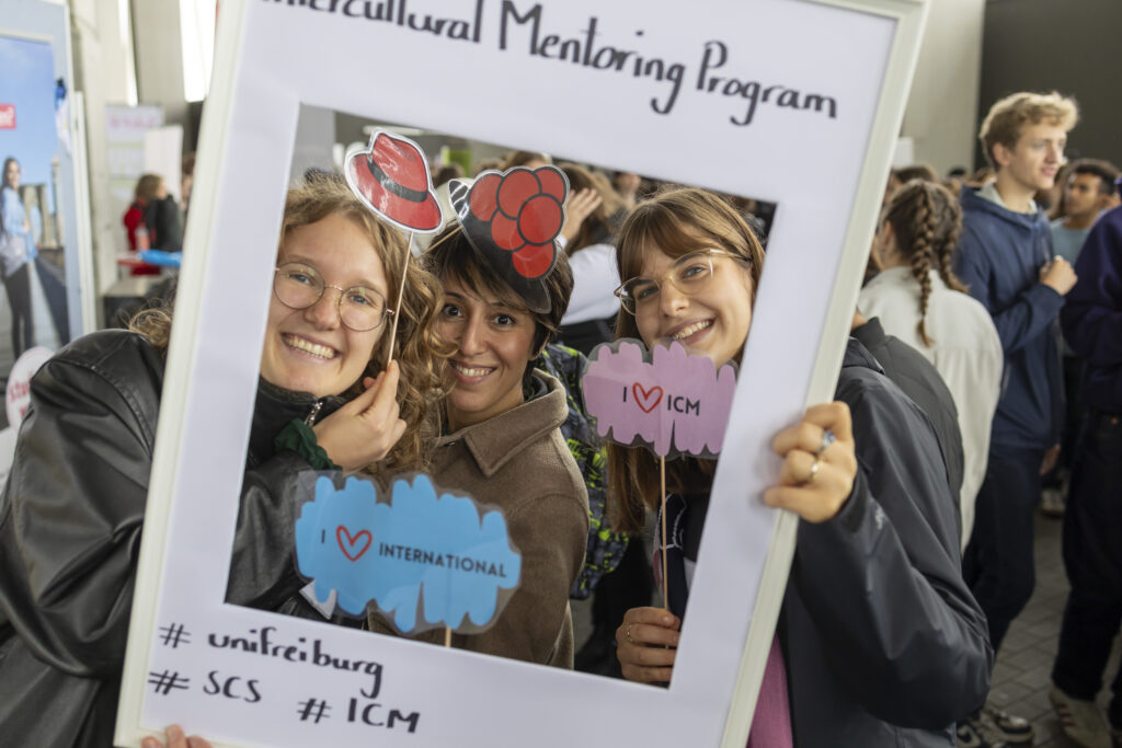 Studentinnen lachen durch einen Rahmen in die Kamera.