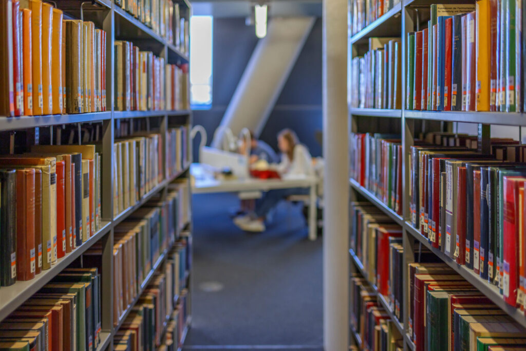 Im Hintergrund sieht unscharf zwei Studierende in der Universitätsbibliothek Freiburg. Im Vordergrund zwei Regalreihen mit Büchern, die auf die beiden zulaufen,