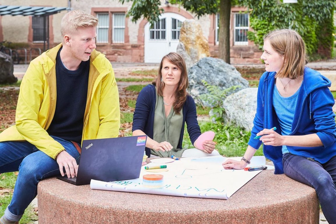 Zwei Studierende und die Nachhaltigkeitsmanagerin sitzen im Freien und konzipieren neue Aktivitäten im Bereich Nachhaltigkeit.