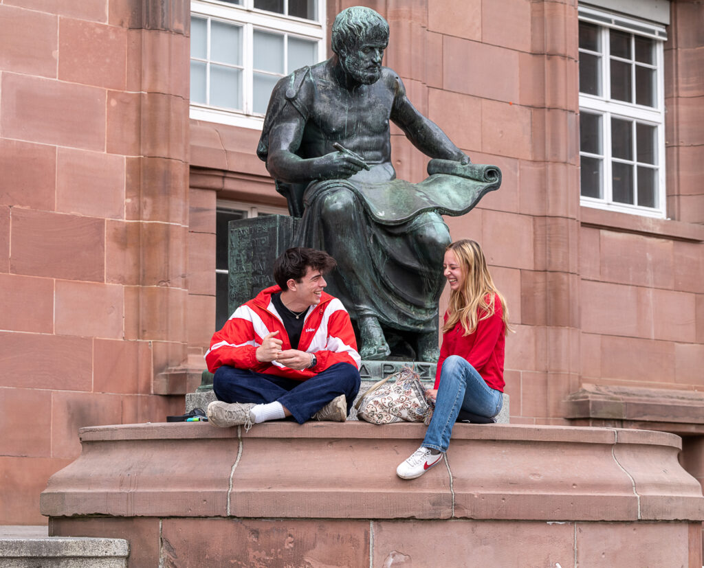 Zwei junge Menschen sitzen lachend unterhalb einer Statue