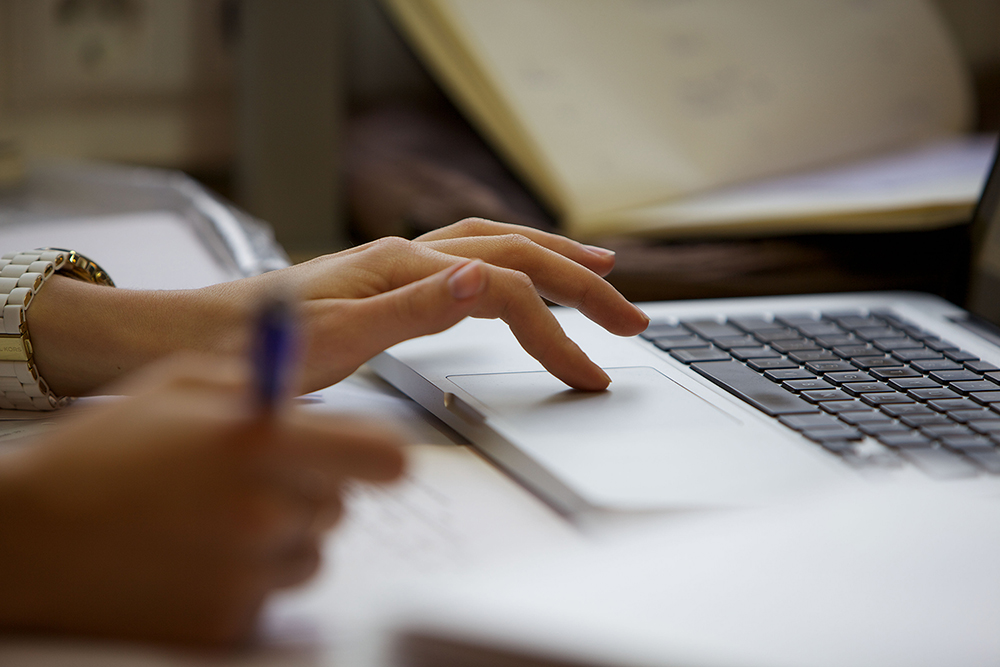 Eine Nahaufnahme einer Hand, die das Touchpad eines Laptops bedient. Die zweite Hand schreibt.