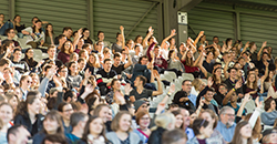 Hunderte Studierende sitzen in einem Fußballstadion. Sie sind fröhlich und lachen, einige strecken die Hände in die Höhe.