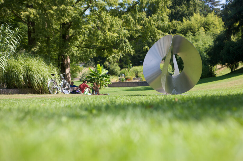 Studierende sitzen bei sommerlichem Wetter auf der Wiese im Botanischen Garten und im Vordergrund befindet sich eine moderne Skulptur aus Metall