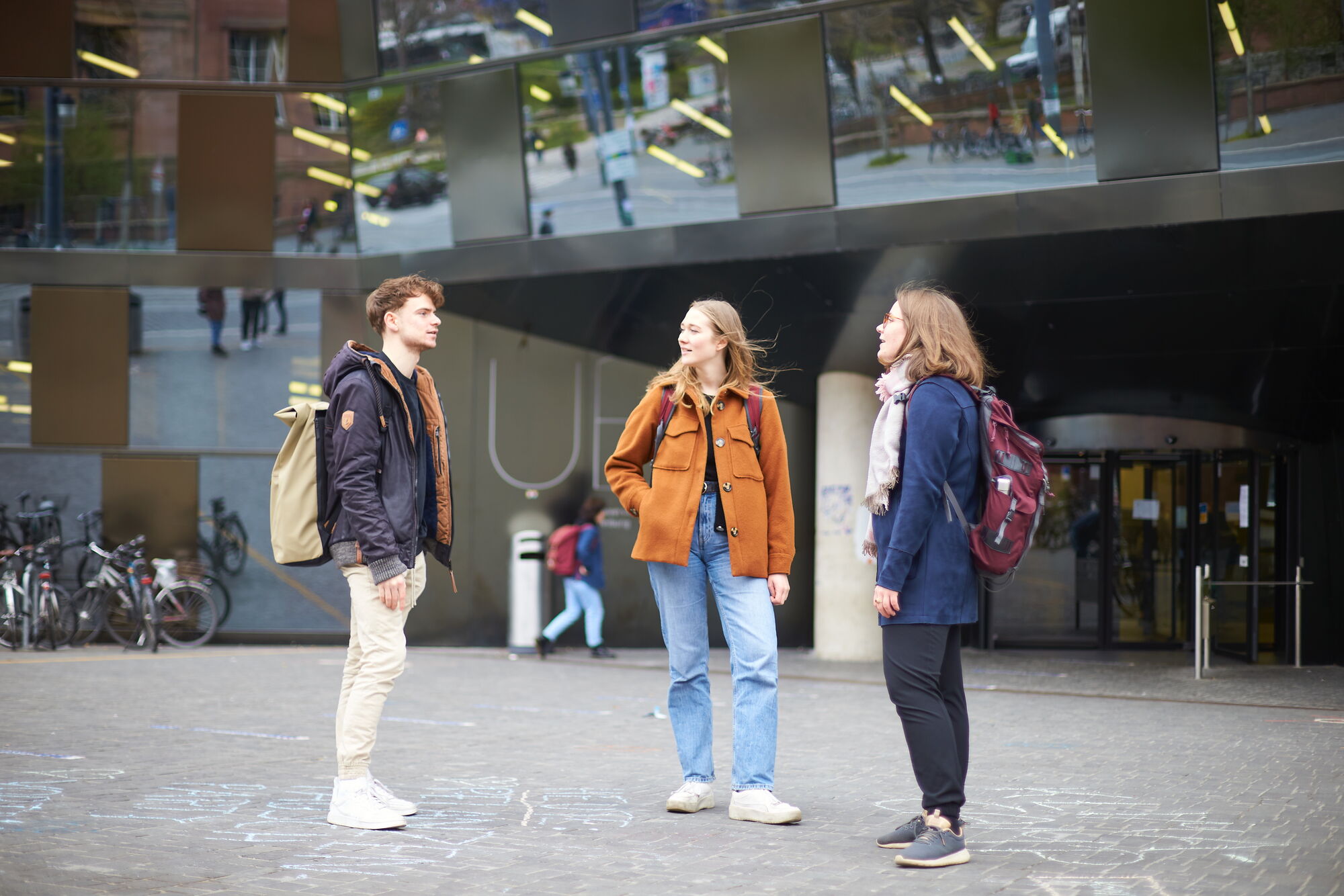 Drei Studierende stehn vor der UB und unterhalten sich. Sie haben Wintermäntel an