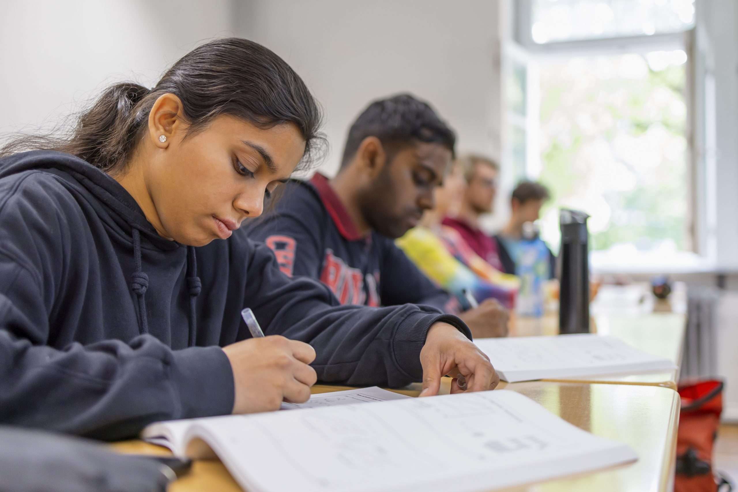 Studierende sitzen in einem Seminarraum. Eine junge Frau im Vordergrund schreibt in ihre geöffneten Unterlagen.