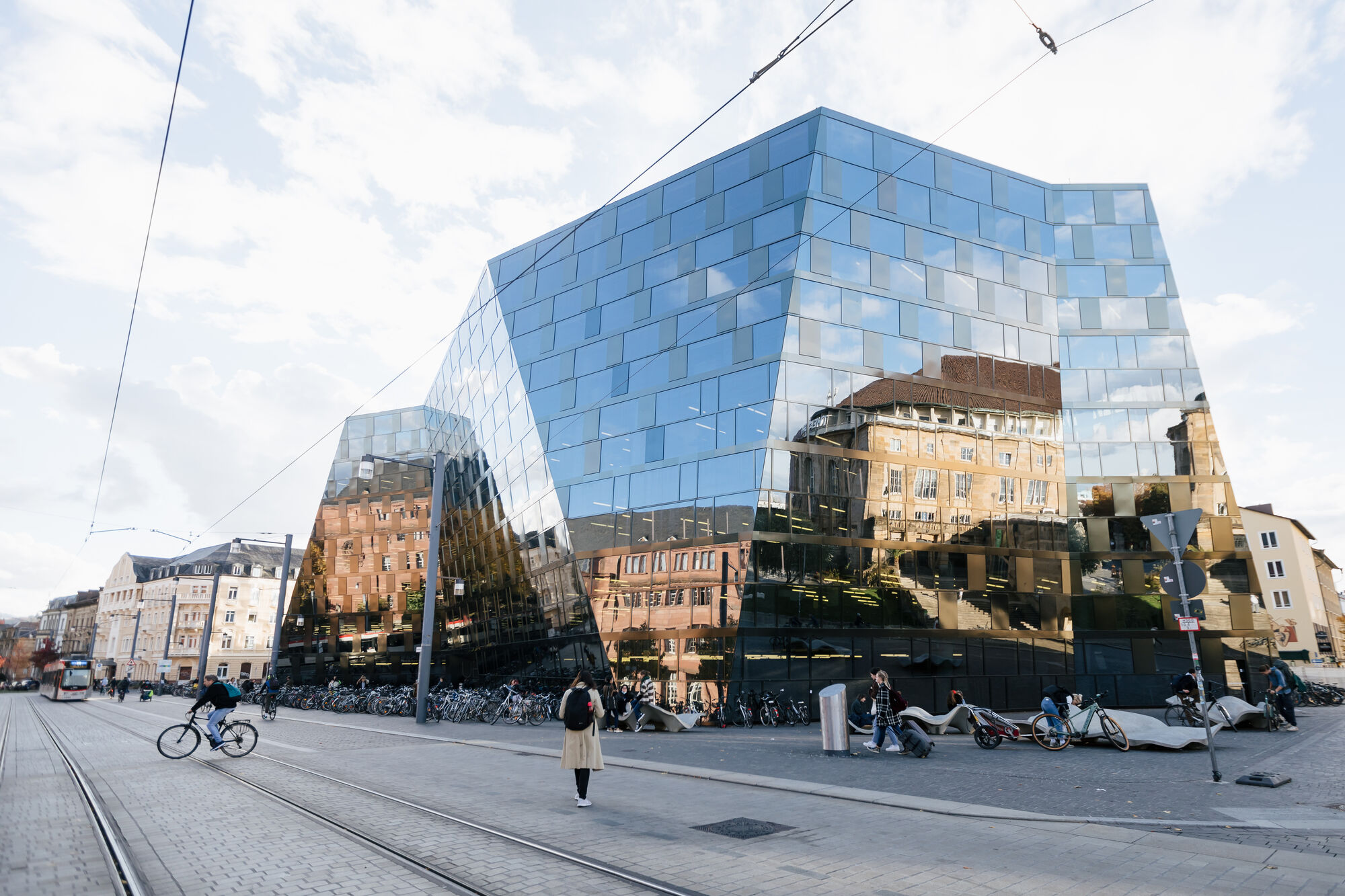 Die Fassade der Universitätsbibliothek, in der sich KGI und Theater spiegeln. Eine Straßenbahn fährt an, ein Radfahrer überquert die Straße, eine Person blickt in Richtung UB.